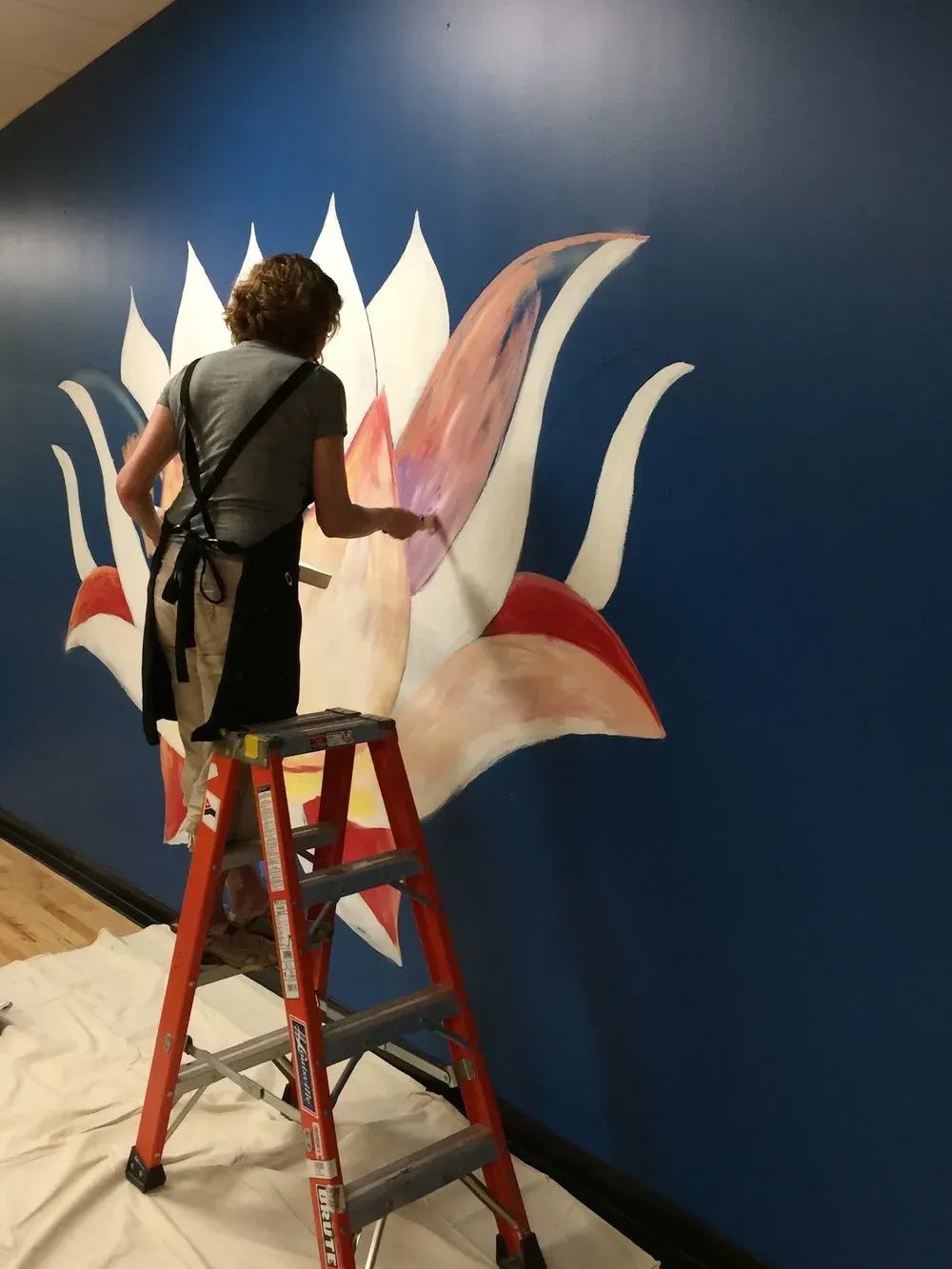 Person on a ladder painting a mural of a lotus flower on a blue wall.