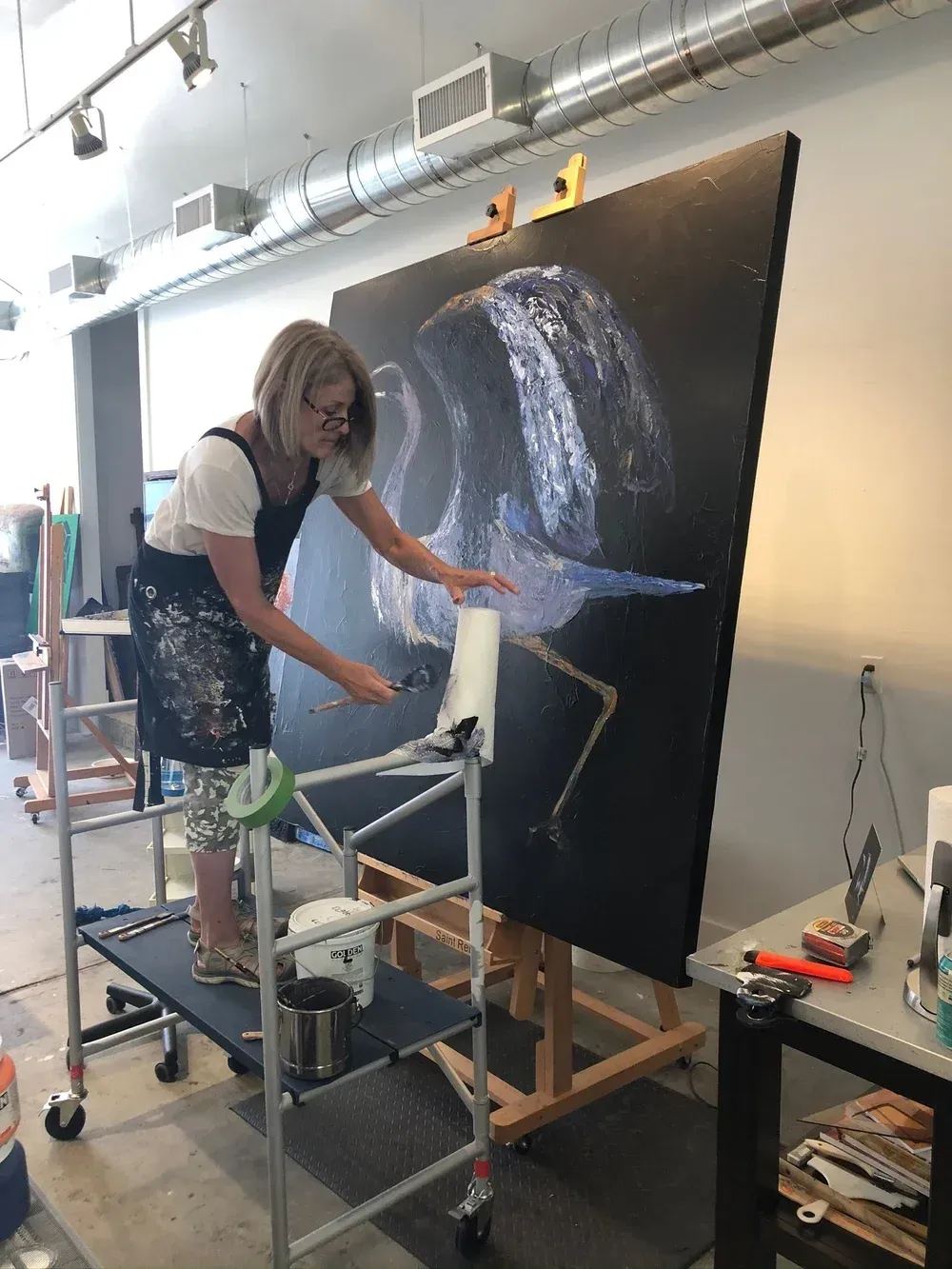 Woman painting a large blue bird on a canvas in an art studio, standing on a rolling platform.