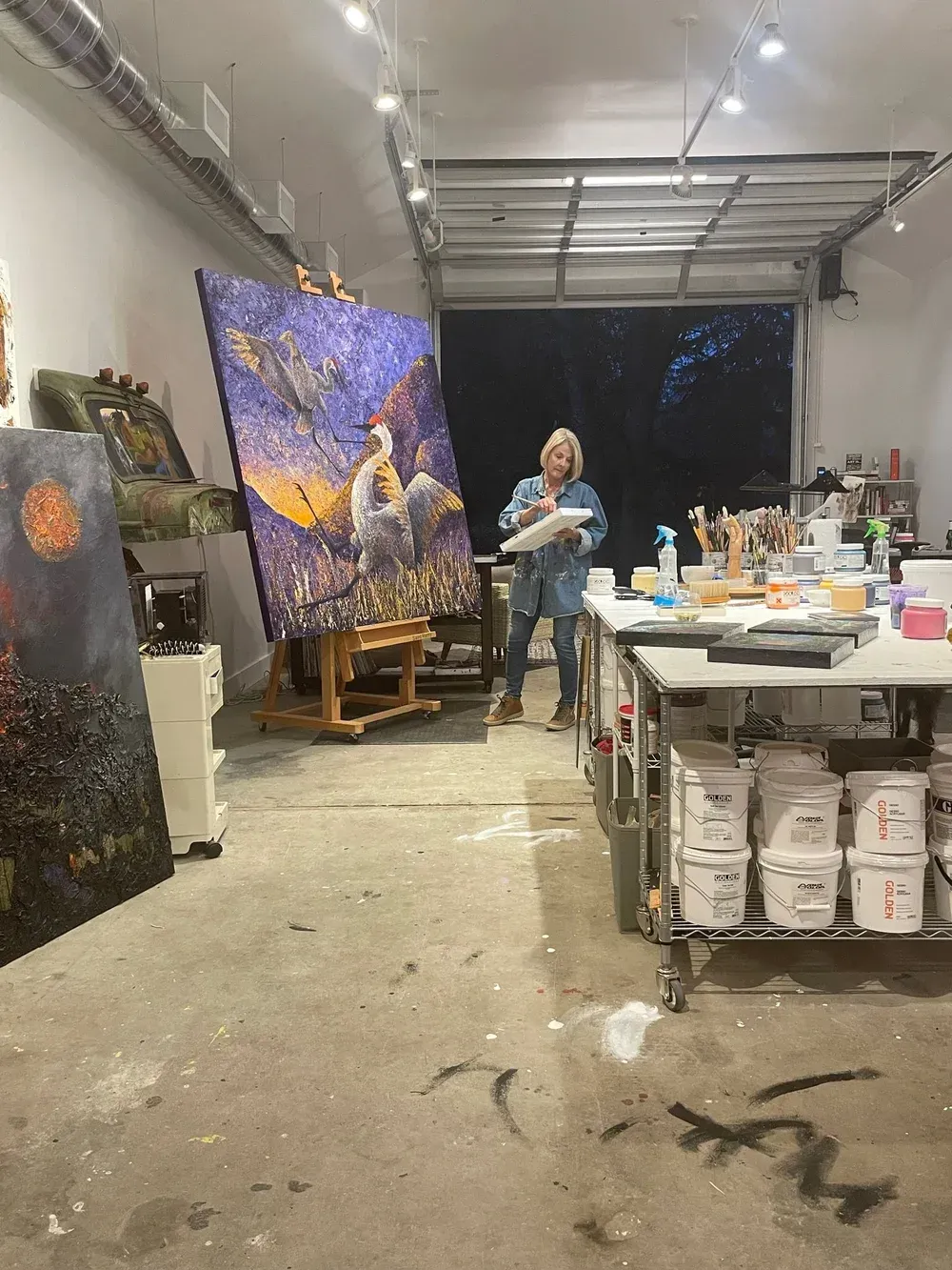 Woman in art studio painting large canvas on easel; work table with art supplies.