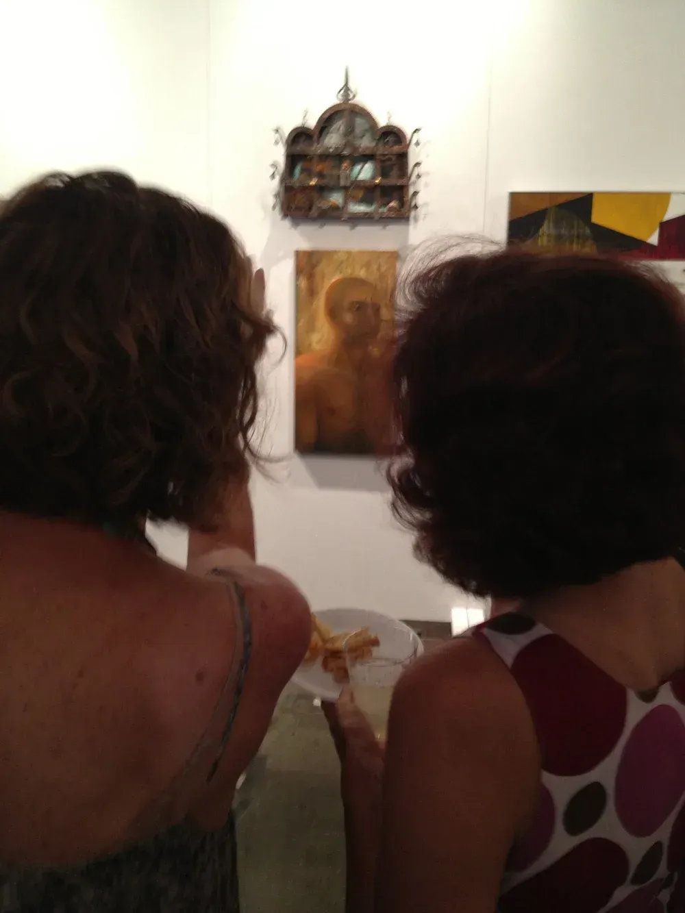 Two people view art at a gallery, one holding a plate. A painting of a figure is on the wall.