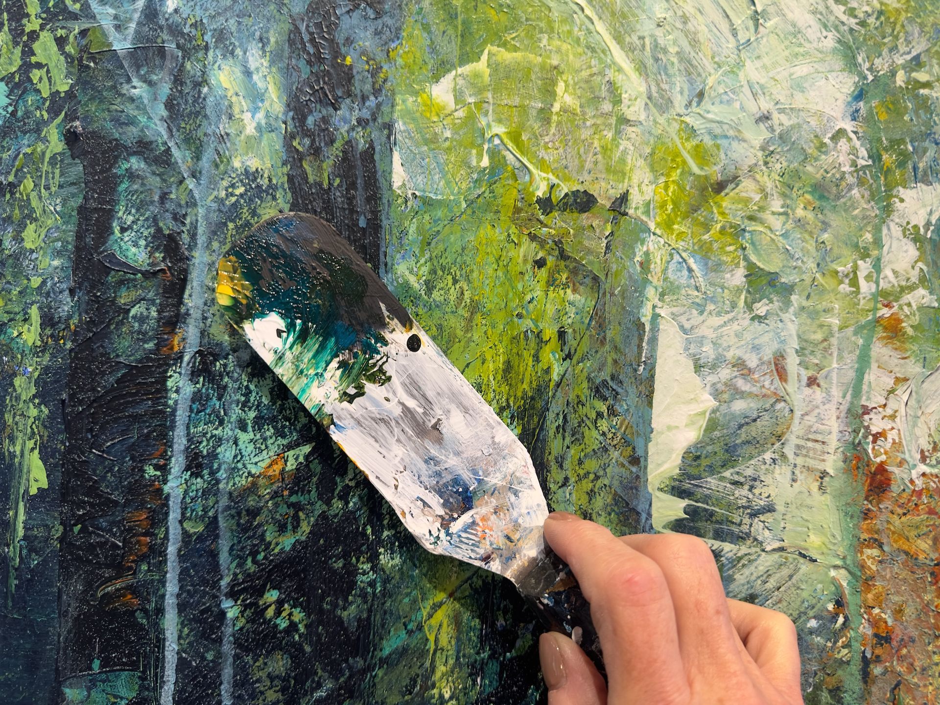 Hand using a palette knife to paint a textured abstract artwork in shades of green, blue, and yellow.