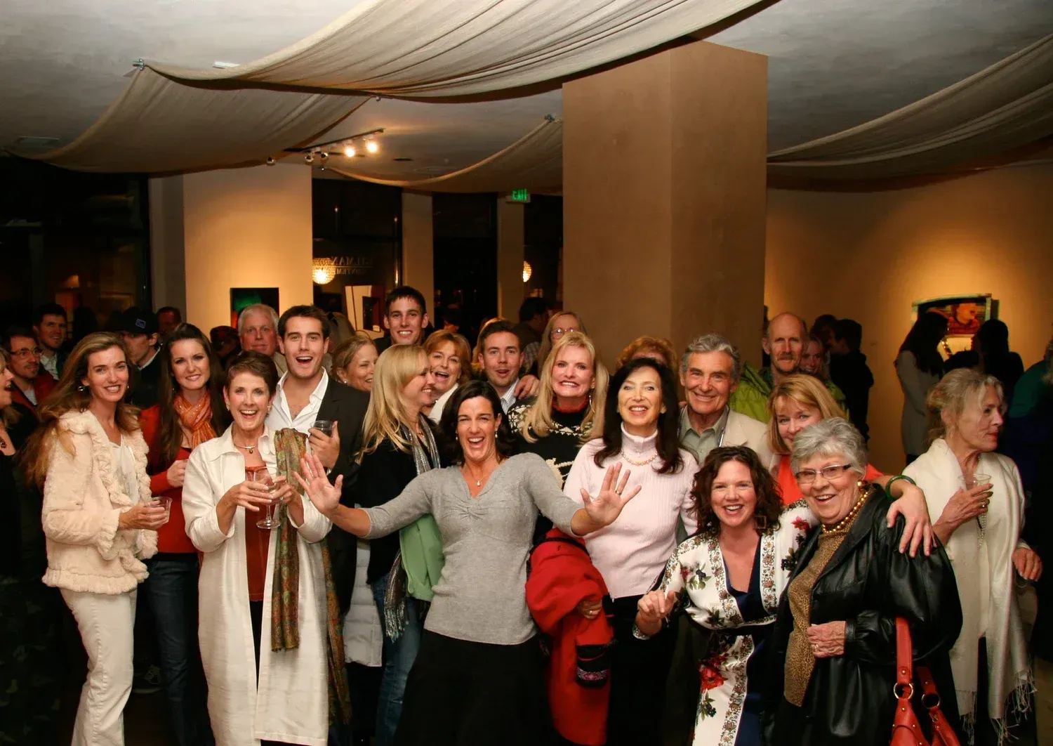 Group of people at an event, some smiling and gesturing towards the camera, indoor setting with artwork.