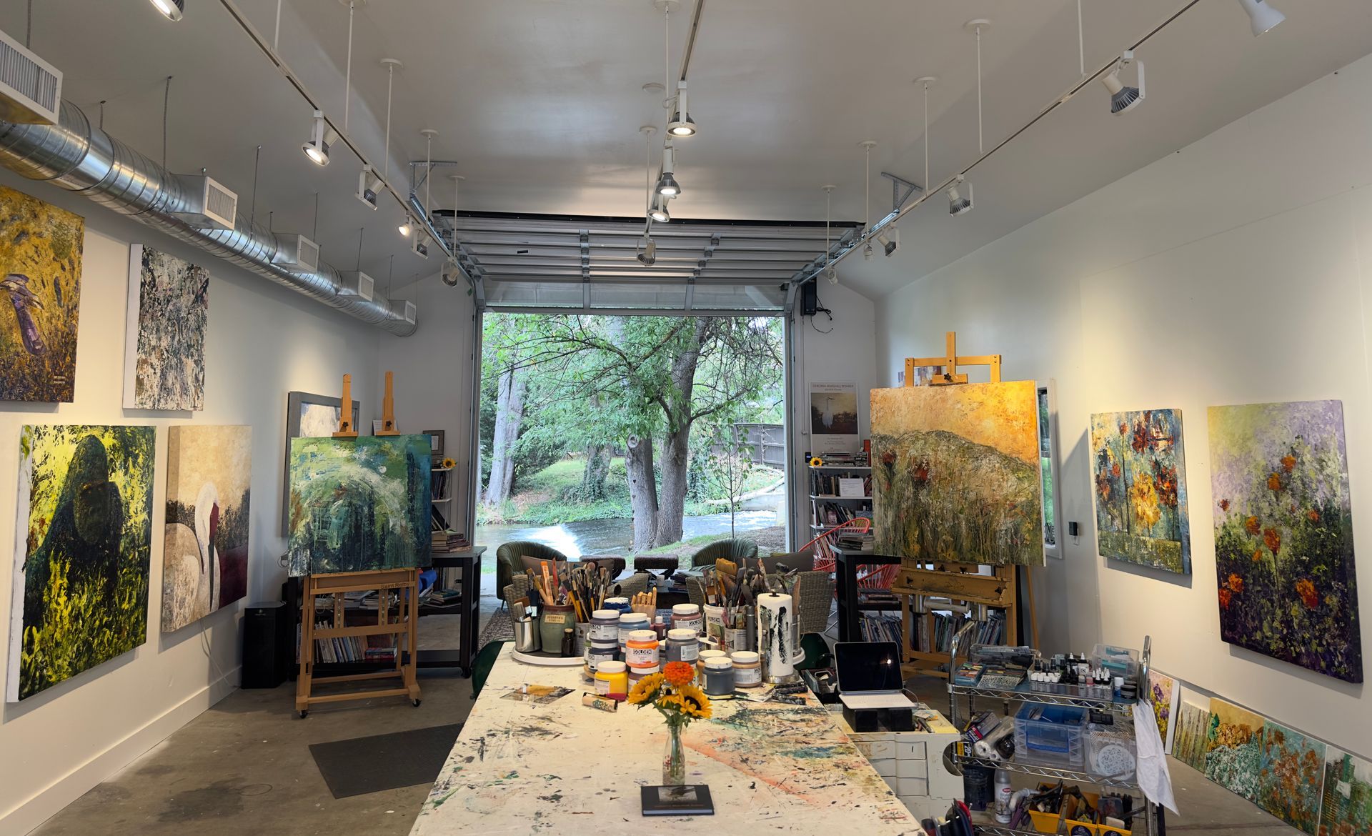 An art studio with paintings on the walls, a long table covered in art supplies, and a large open doorway with trees visible.