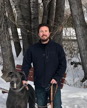 Man in blue jacket stands with a Great Dane dog in a snowy park setting.