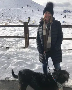 Woman in winter coat with black dog in snowy landscape.