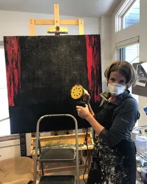 Woman sanding large black and red painting on an easel, wearing a respirator mask, indoors.