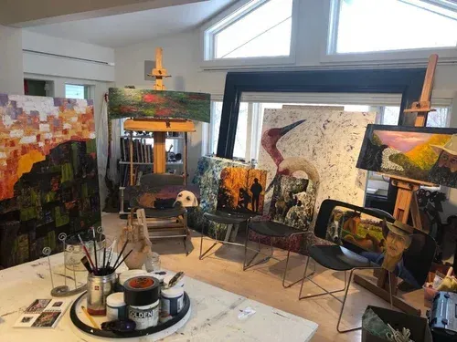 Artist's studio with paintings on easels. Paint supplies on a table. Bright room with natural light.