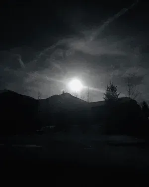 Silhouetted mountain and trees under a bright moon in a cloudy, dark sky.