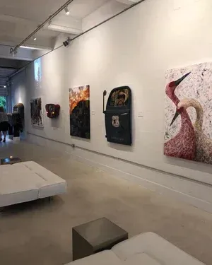 Art gallery with paintings and sculptures on white walls. Includes a person, a mailbox art piece, and white benches.