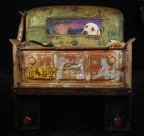 Rusty Ford truck bed with a yellow lab looking out the window. Tools in the bed, license plate, black backdrop.