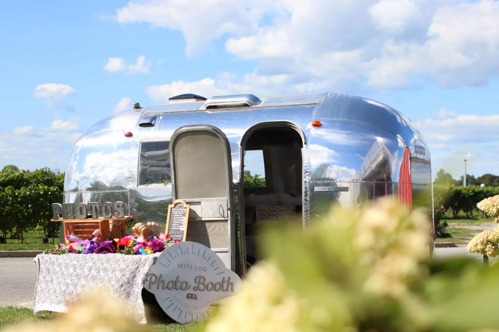 Shiny silver photo booth trailer set up outdoors under a blue sky, with flowers in the foreground.