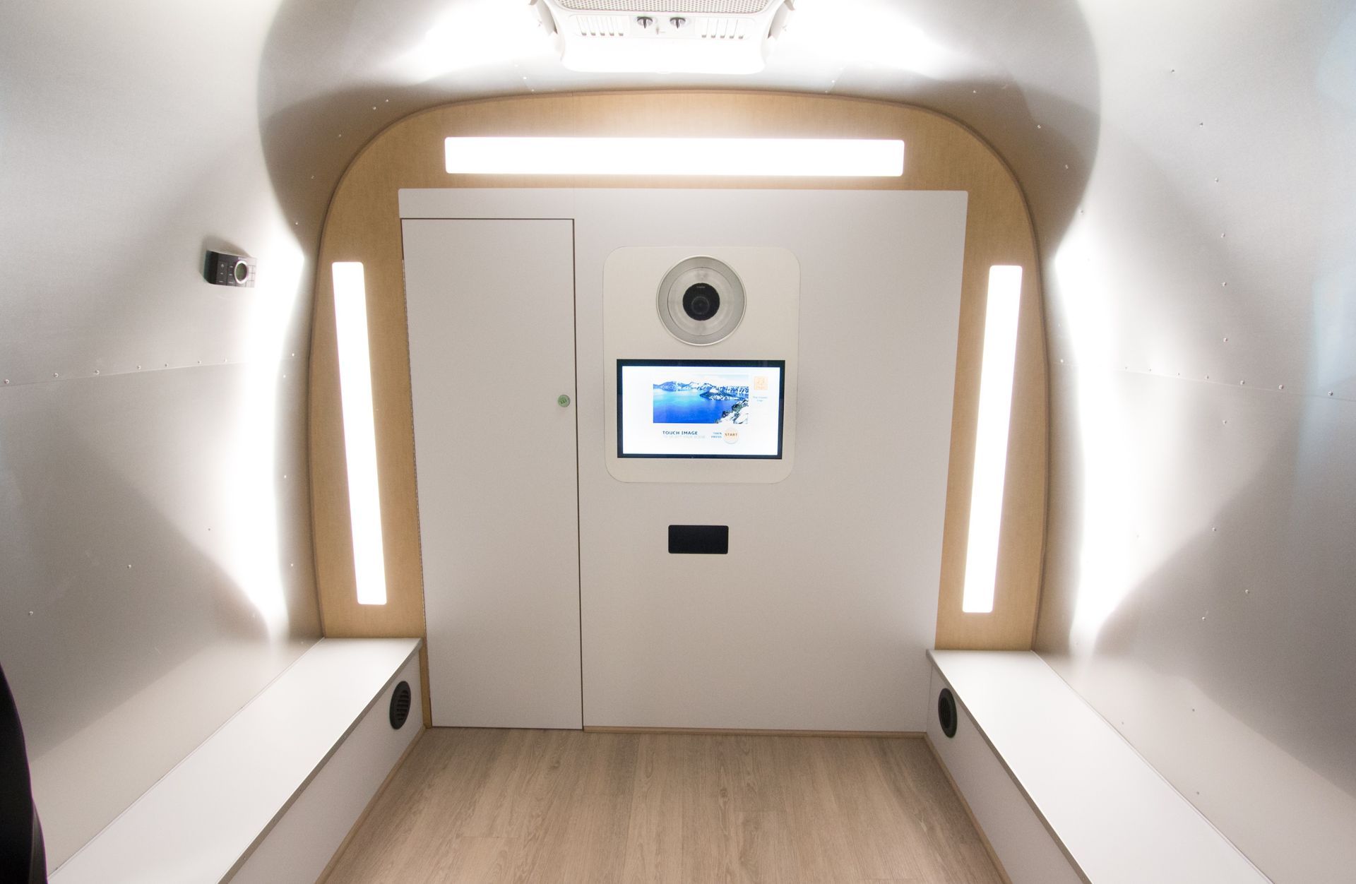 Interior of a white pod with benches, a door, screen, and lights, possibly for a medical scan.