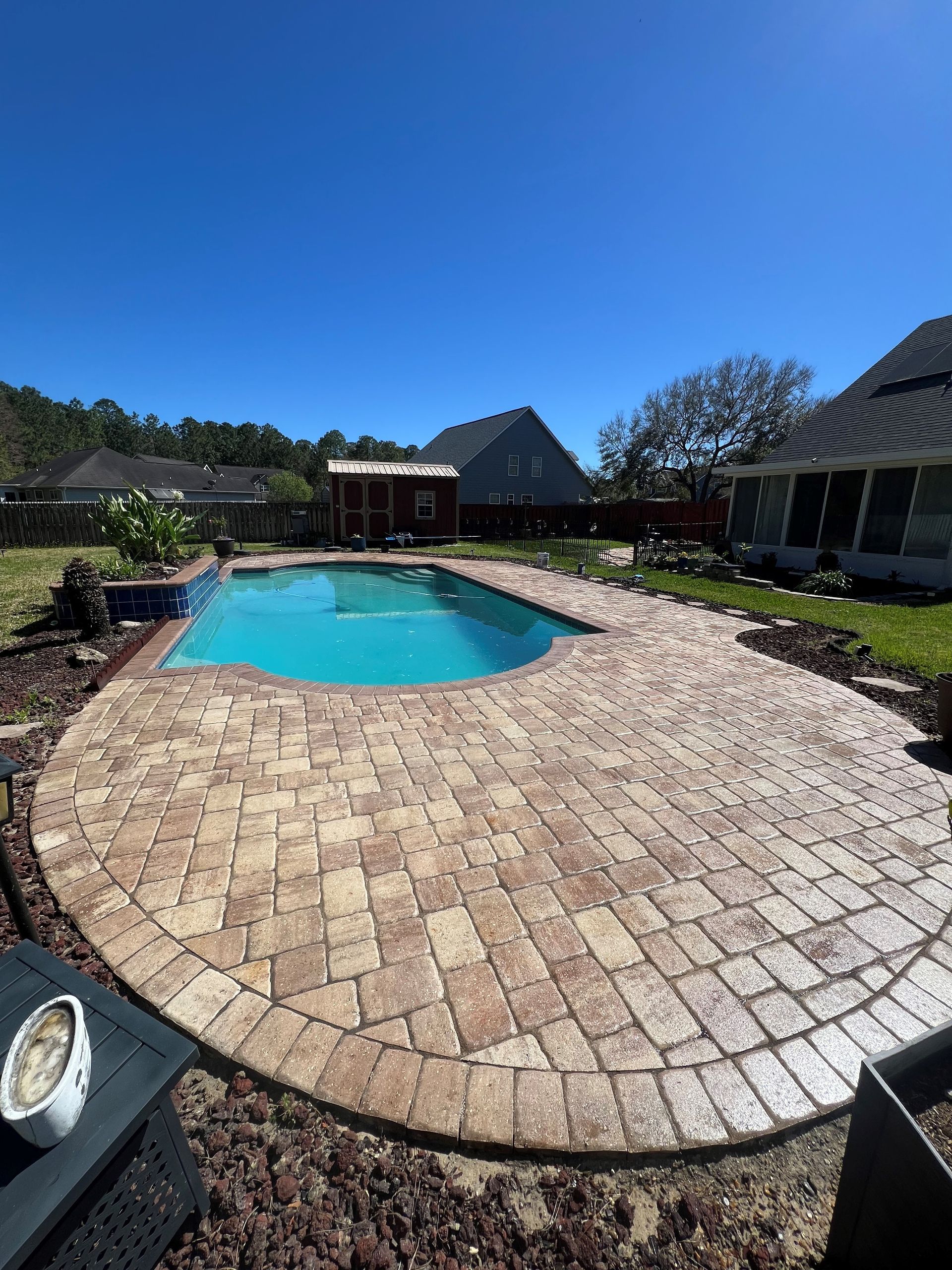 A brick patio surrounds a blue swimming pool on a sunny day.