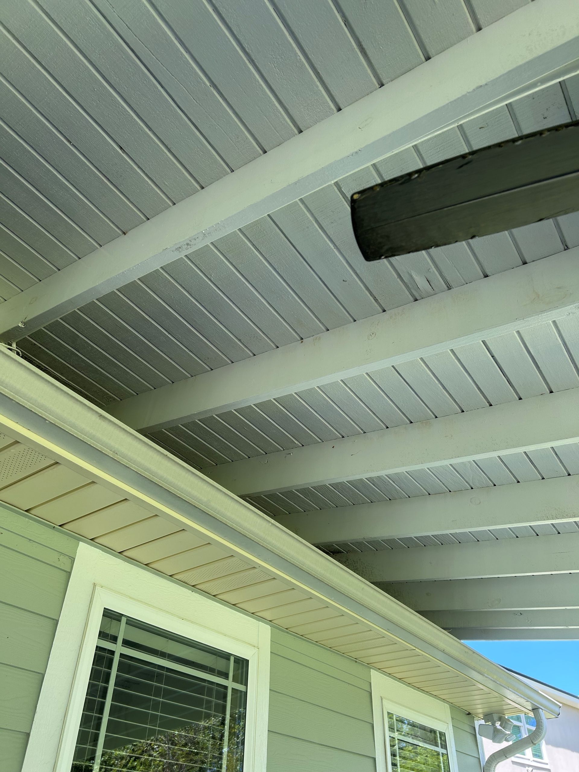 View of a light gray porch ceiling with horizontal planks and white beams, adjacent to a light green wall with windows and a gutter.