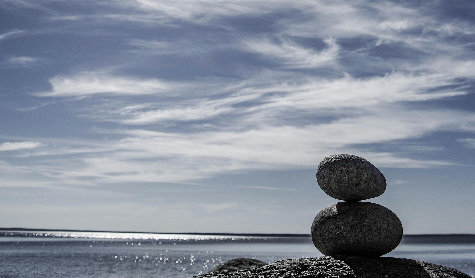 Two stones balanced on a shoreline, set against a bright blue sky with wispy clouds over a calm body of water.