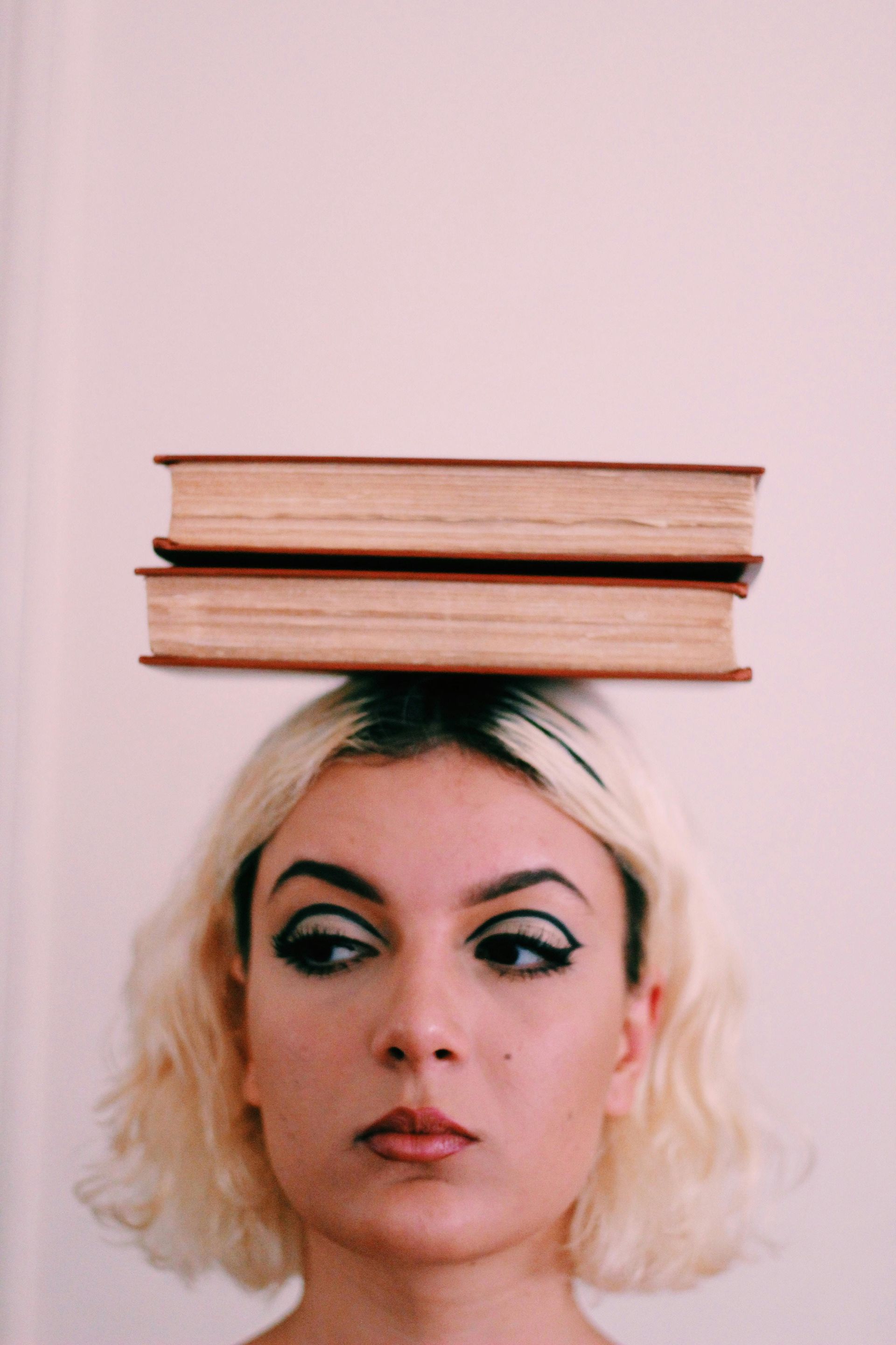 Woman with blonde hair balancing two books on her head, looking to the side with a thoughtful expression.