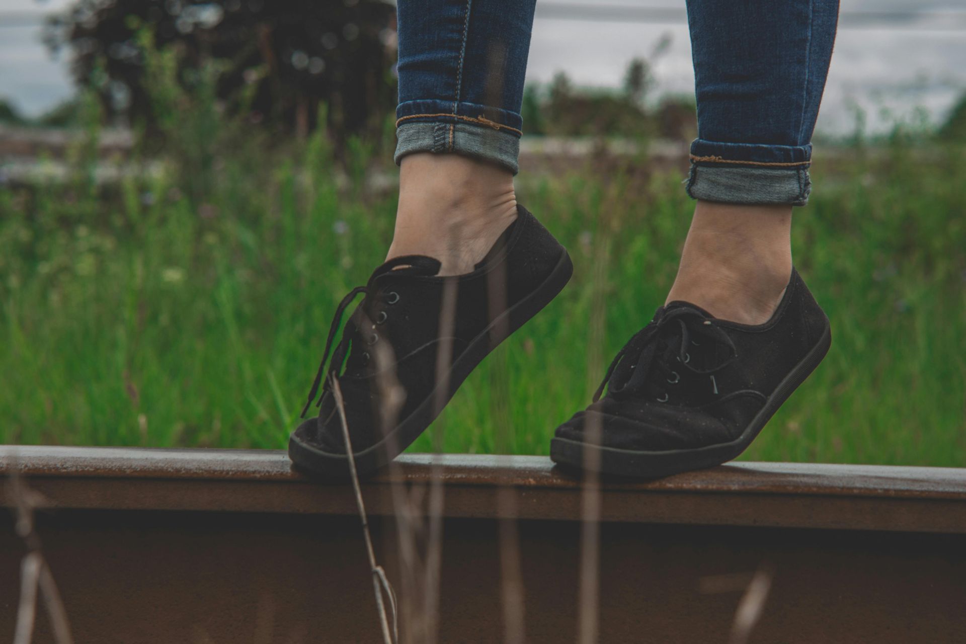 Person's feet in black shoes standing on a train track, wearing blue jeans with a blurred green background.