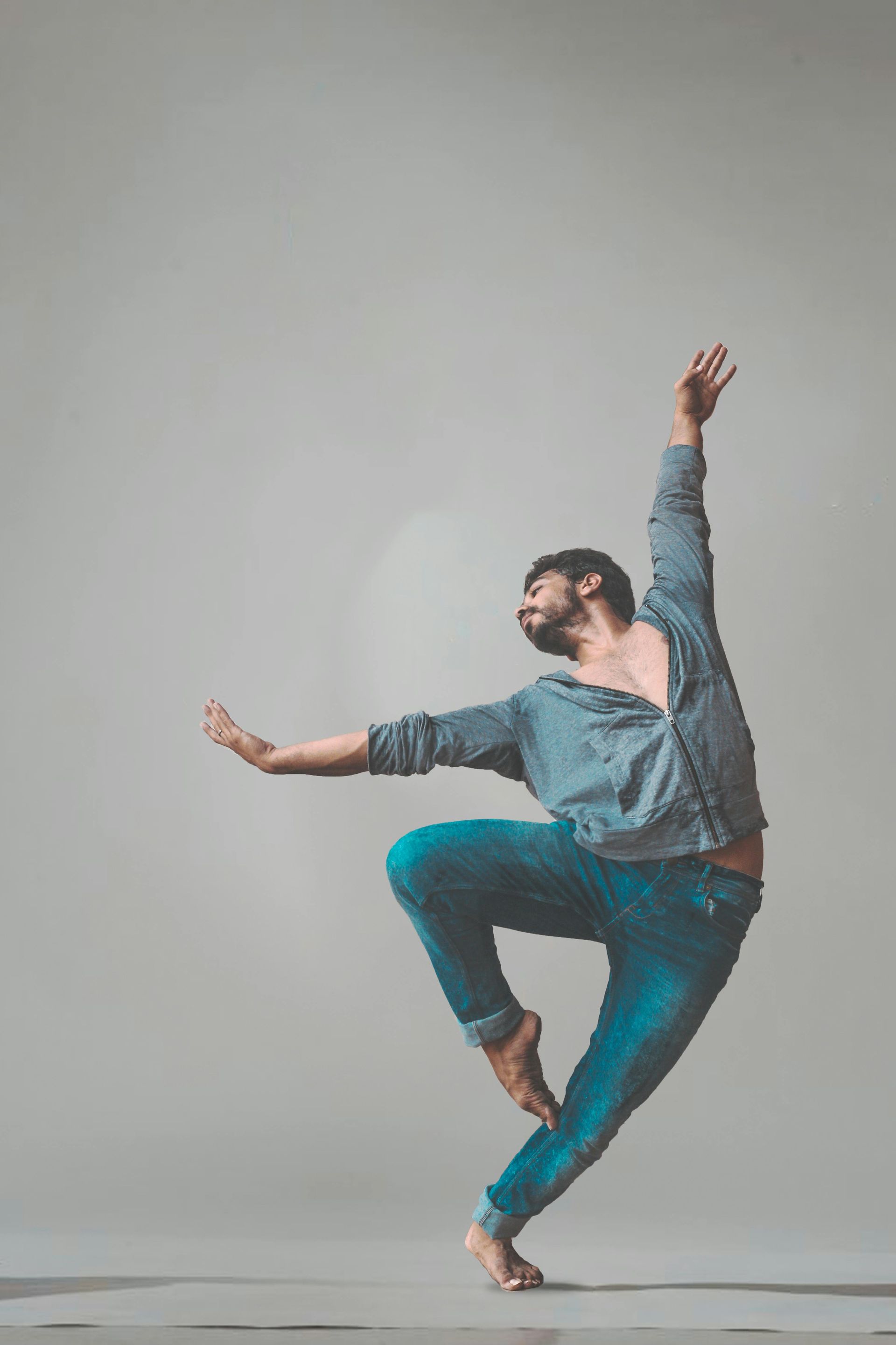 Man dances barefoot, wearing jeans and a blue shirt. He leaps with one arm raised and the other out. Light background.