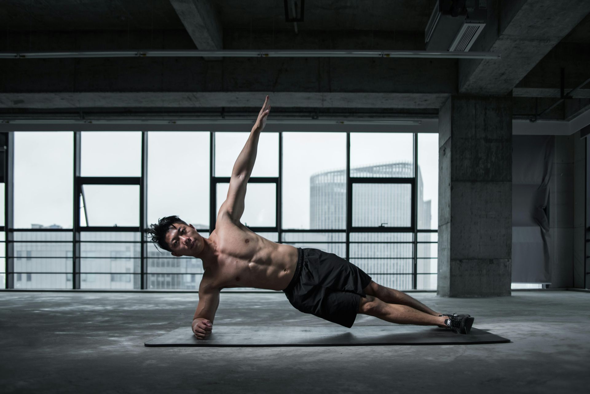Man in a side plank pose, lifting arm, working out in a building with city view.