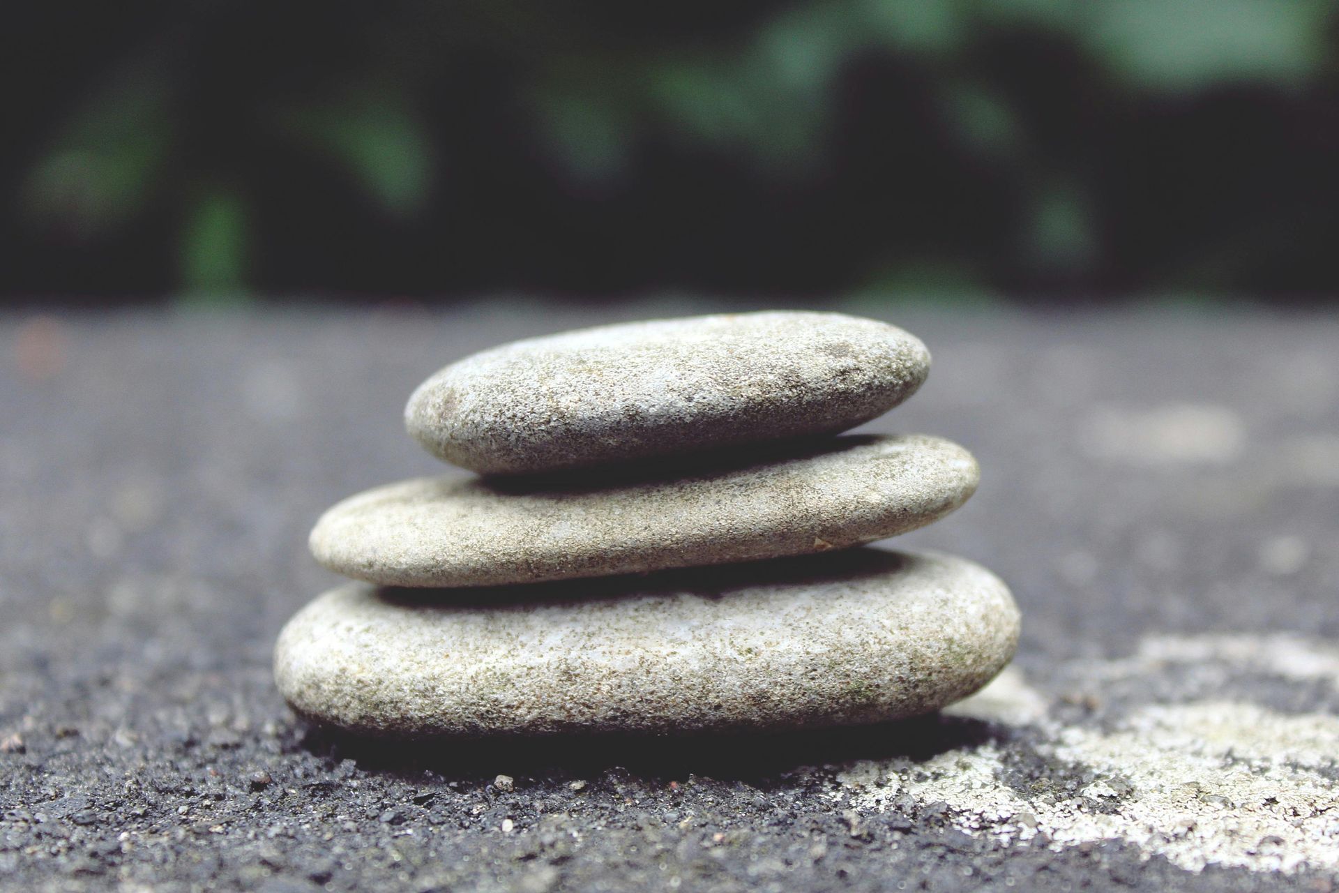 Three gray stones stacked in a balanced tower, outdoors on a dark, textured surface.
