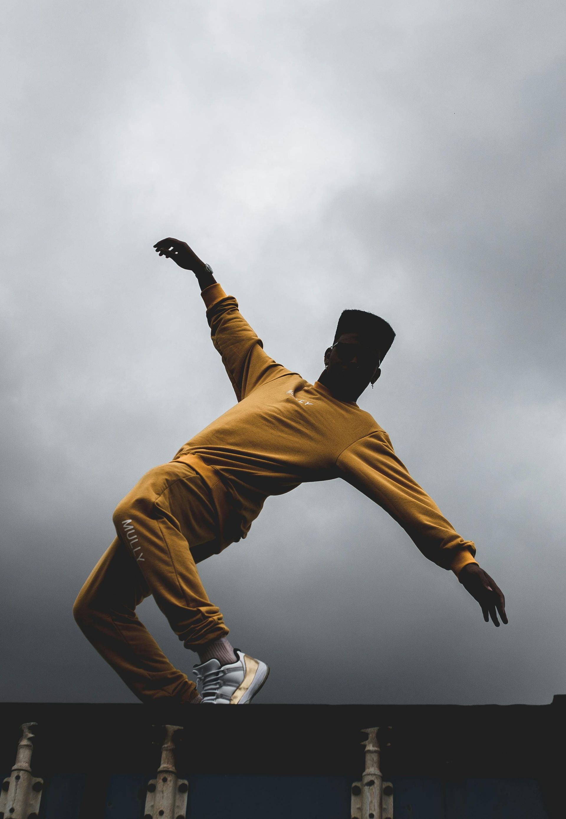 Person in mustard sweatsuit balancing and arching back on a wall, under a stormy sky.