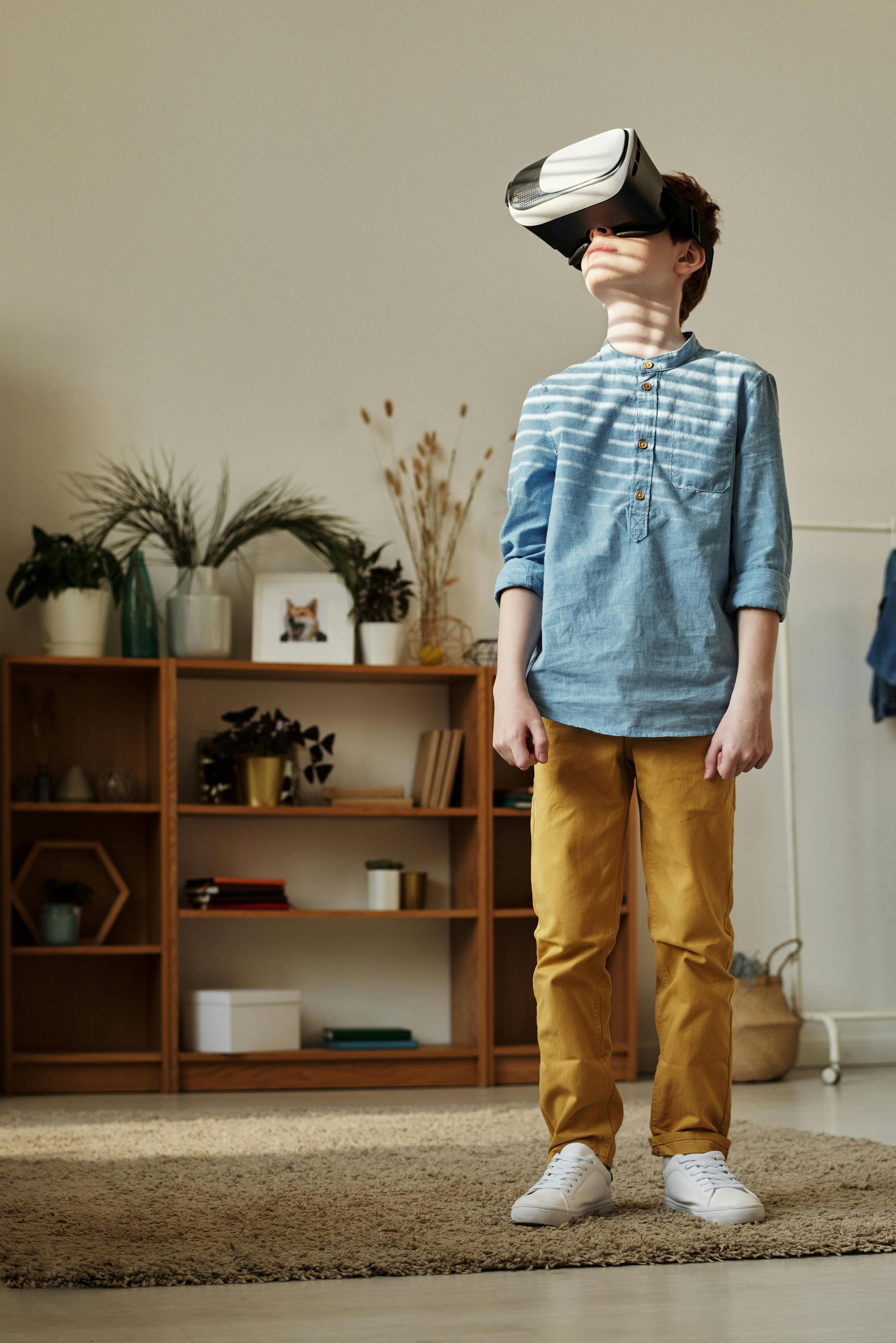 Boy wearing VR headset, looking upwards. Stands in living room with bookshelf, wearing blue shirt and yellow pants.