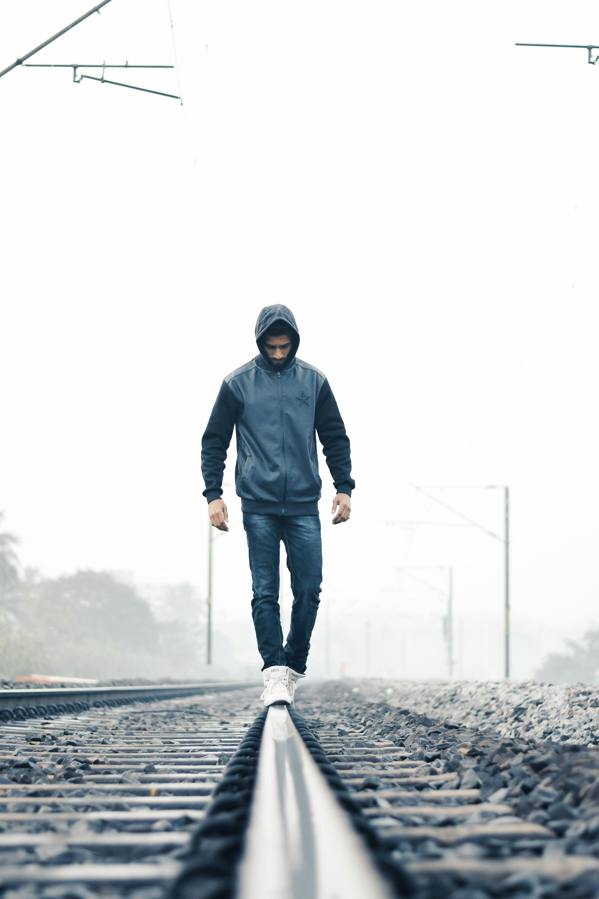 Man in hoodie walks on train tracks in foggy setting.