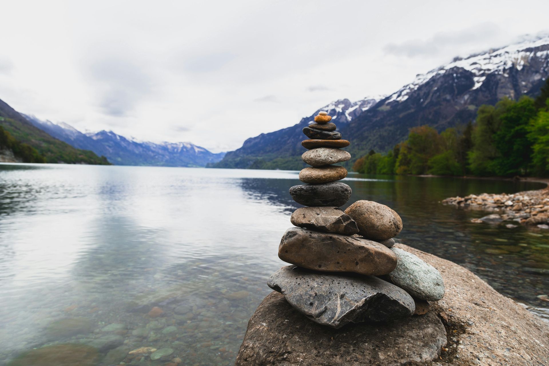 Stone cairn on rock by lake, snow-capped mountains in background, cloudy sky.
