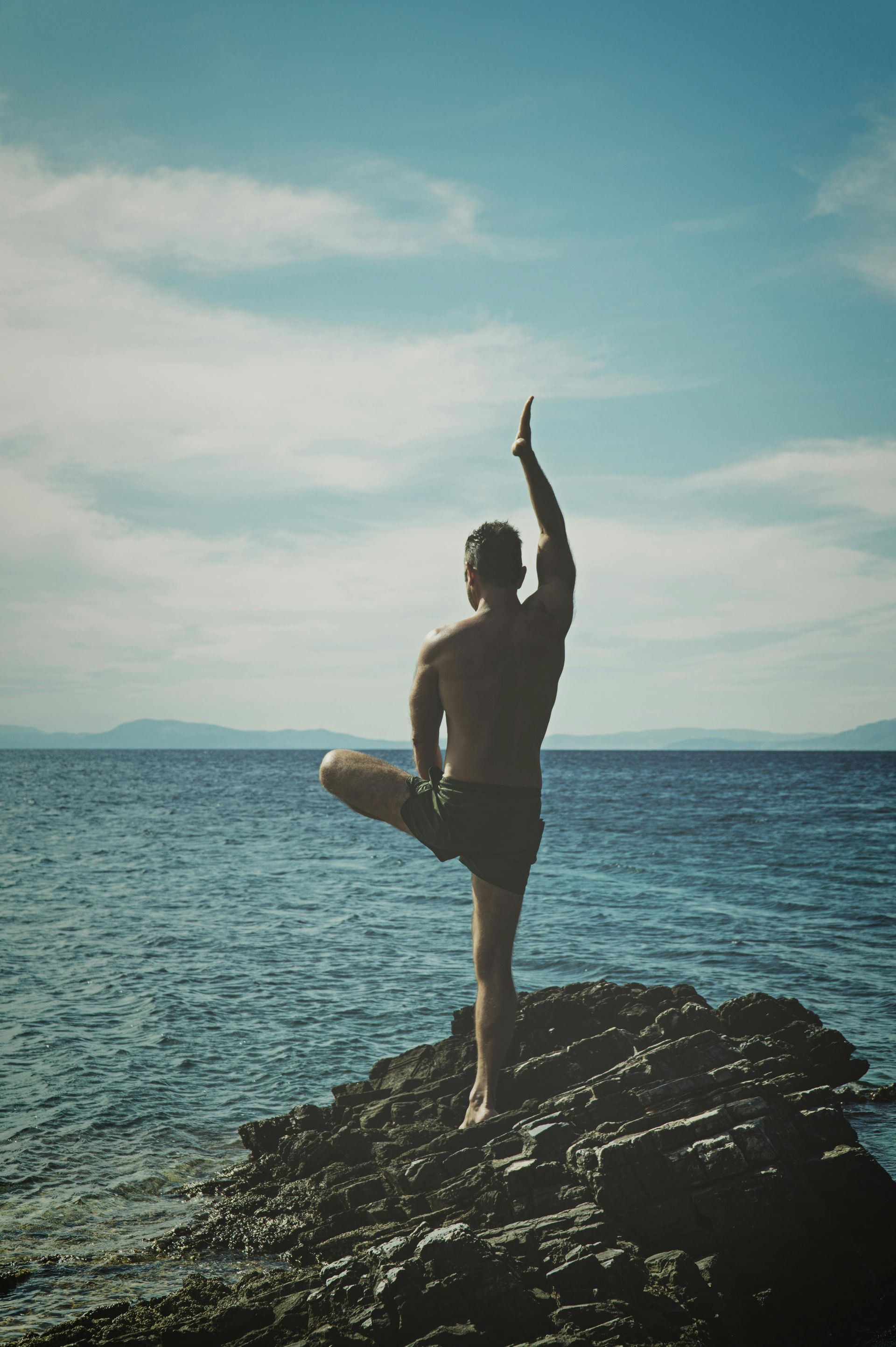 Man balances in a tree pose on a rock by the sea, arms outstretched, blue sky.