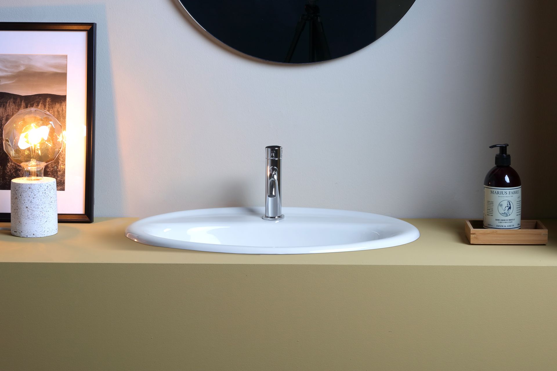 Lavandino ovale bianco con rubinetto cromato, sopra un bancone beige. Flacone di sapone e candela in bella vista.