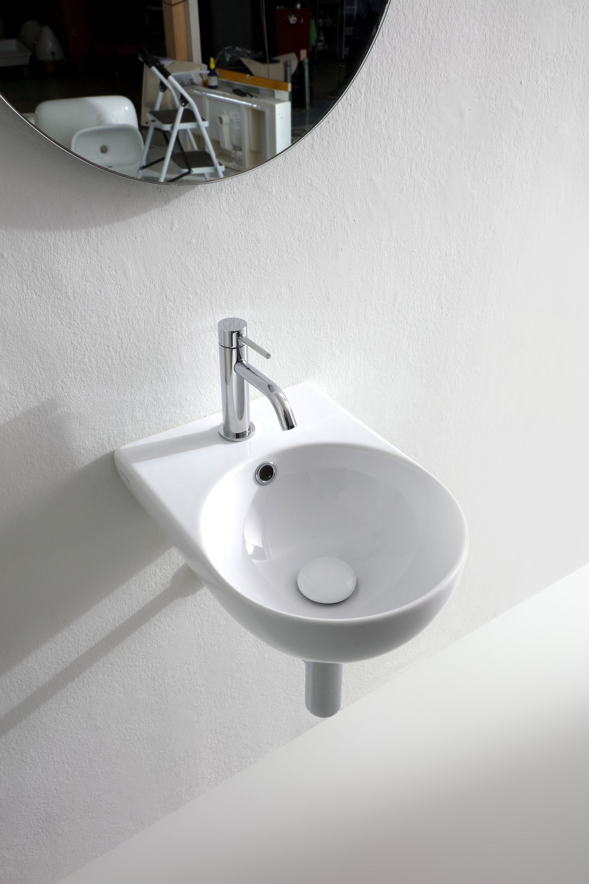 Lavandino bianco curvo a parete con rubinetto e scarico cromati. Specchio riflettente sullo sfondo.