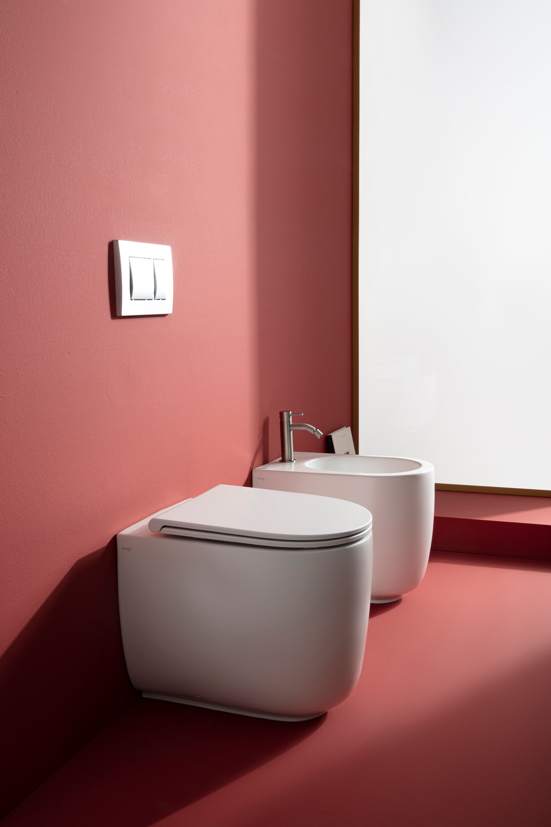Bagno moderno con WC e bidet bianchi contro una parete rossa. Pulsante di scarico argentato.