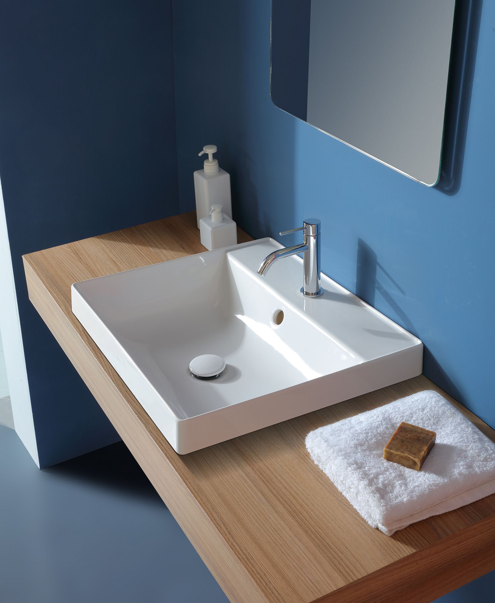 Lavandino rettangolare bianco su un bancone in legno con rubinetto argentato, sapone e asciugamano, contro una parete blu.