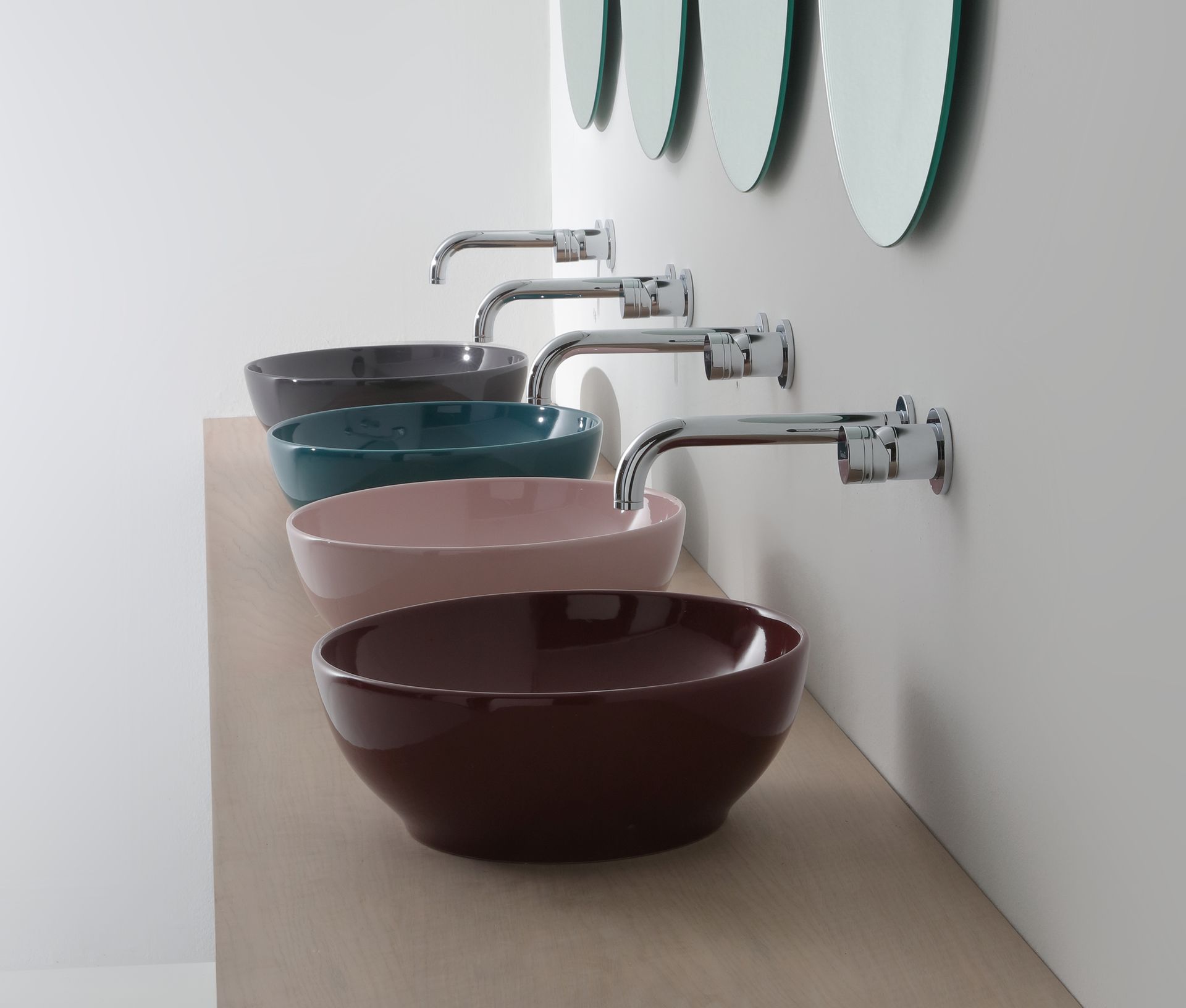 Quattro lavandini colorati con rubinetti a parete su una mensola in legno, sotto specchi rotondi.