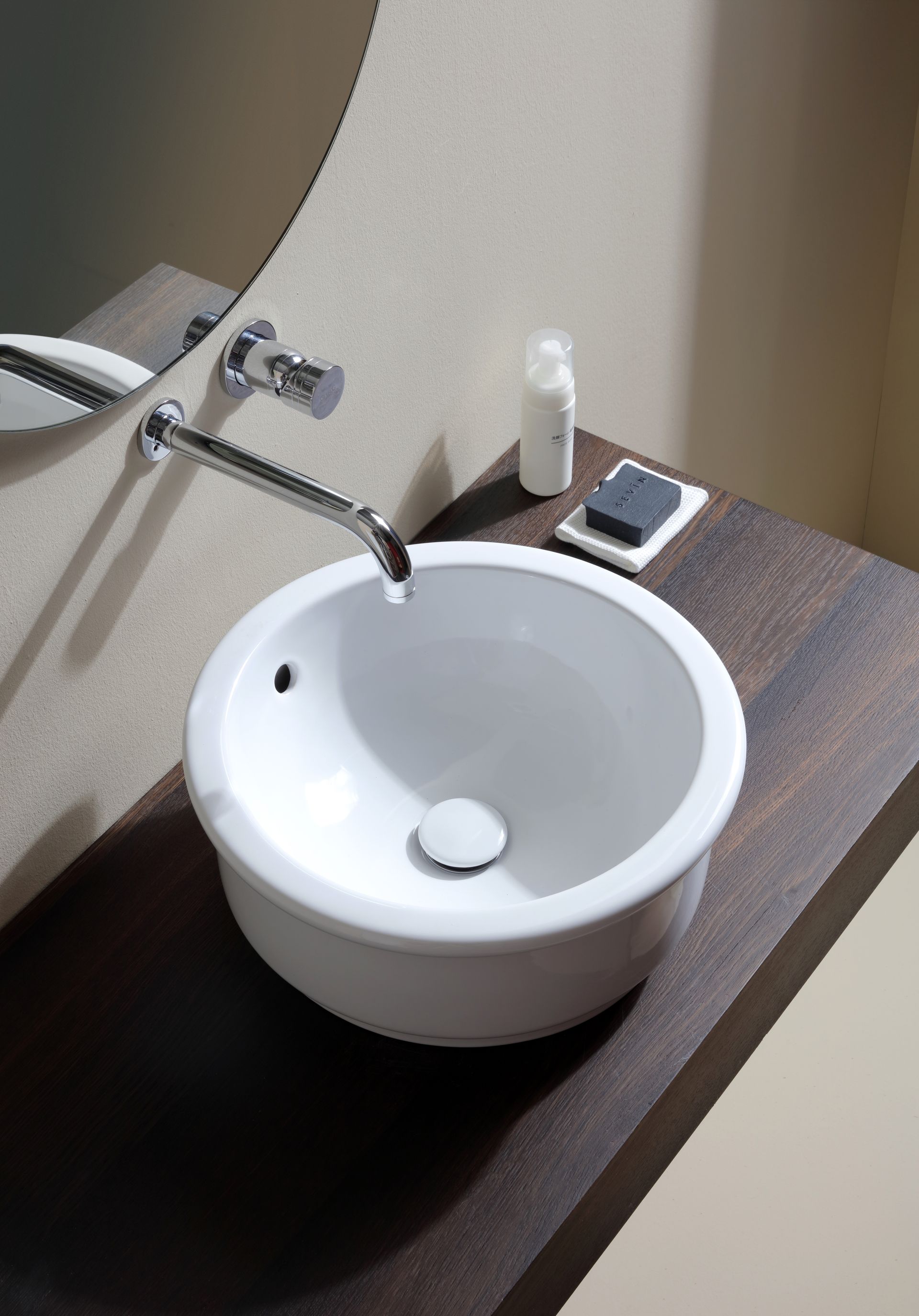 Lavandino circolare bianco su un piano di lavoro in legno scuro con rubinetto cromato e dispenser per il sapone.