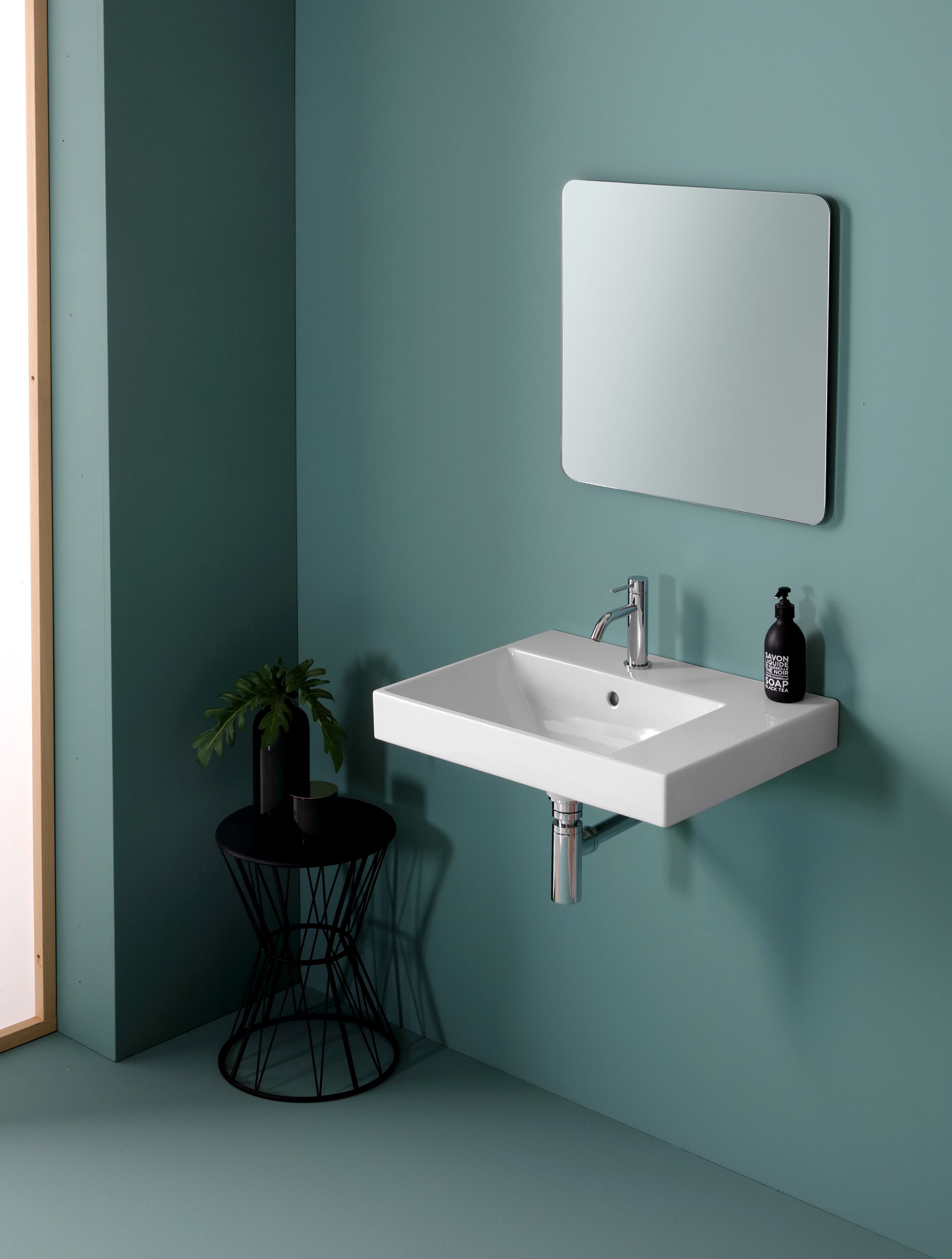 Lavandino bianco da bagno a parete con rubinetto cromato e specchio su parete verde acqua, piccola pianta su tavolino nero.