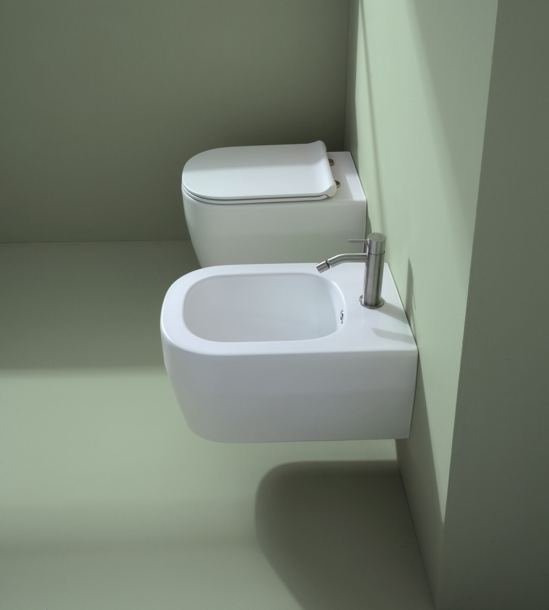 WC e bidet bianchi montati su una parete verde in un bagno. Sul bidet è visibile un rubinetto.