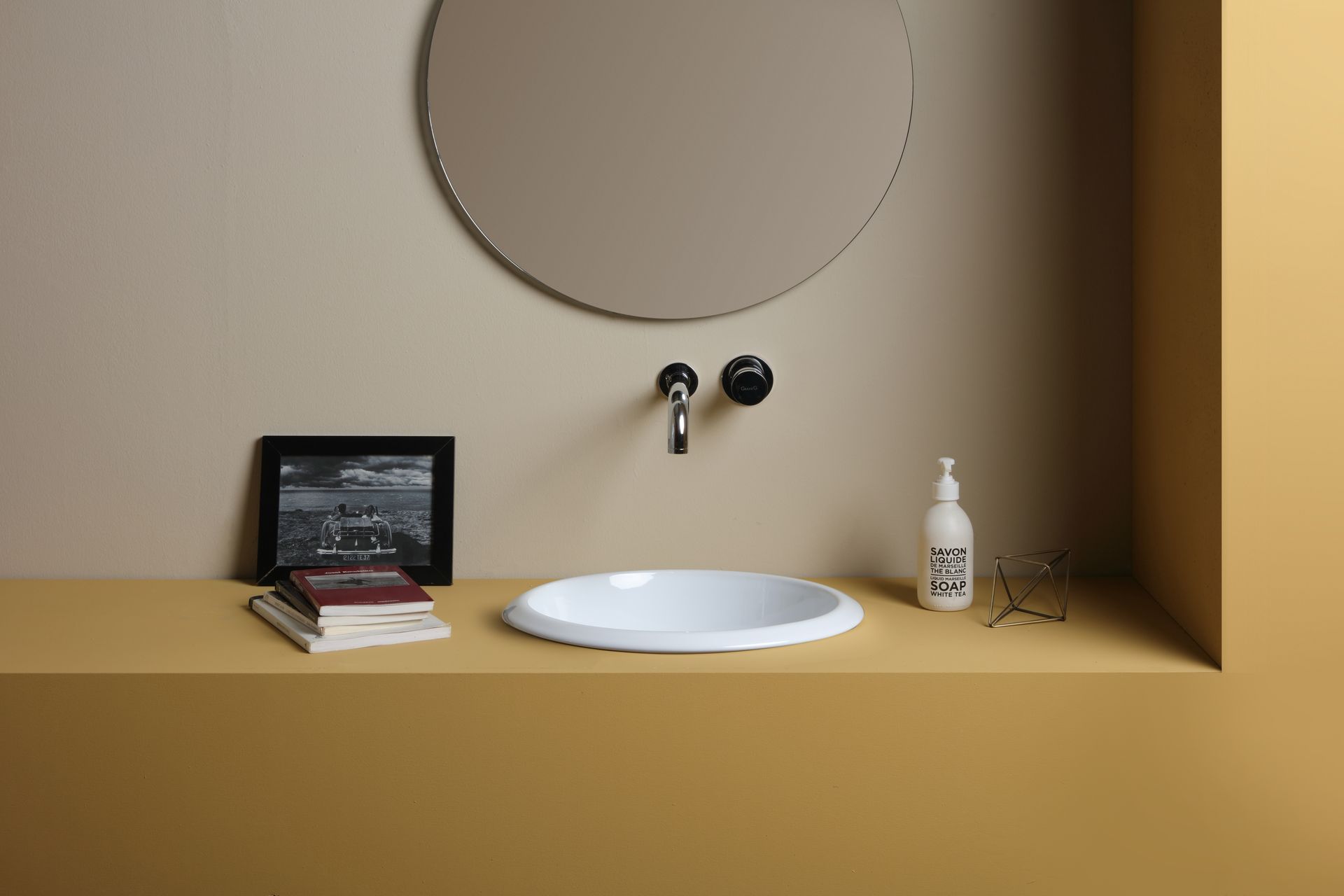 Lavandino bianco con rubinetto cromato, specchio rotondo, pareti beige e bancone giallo.
