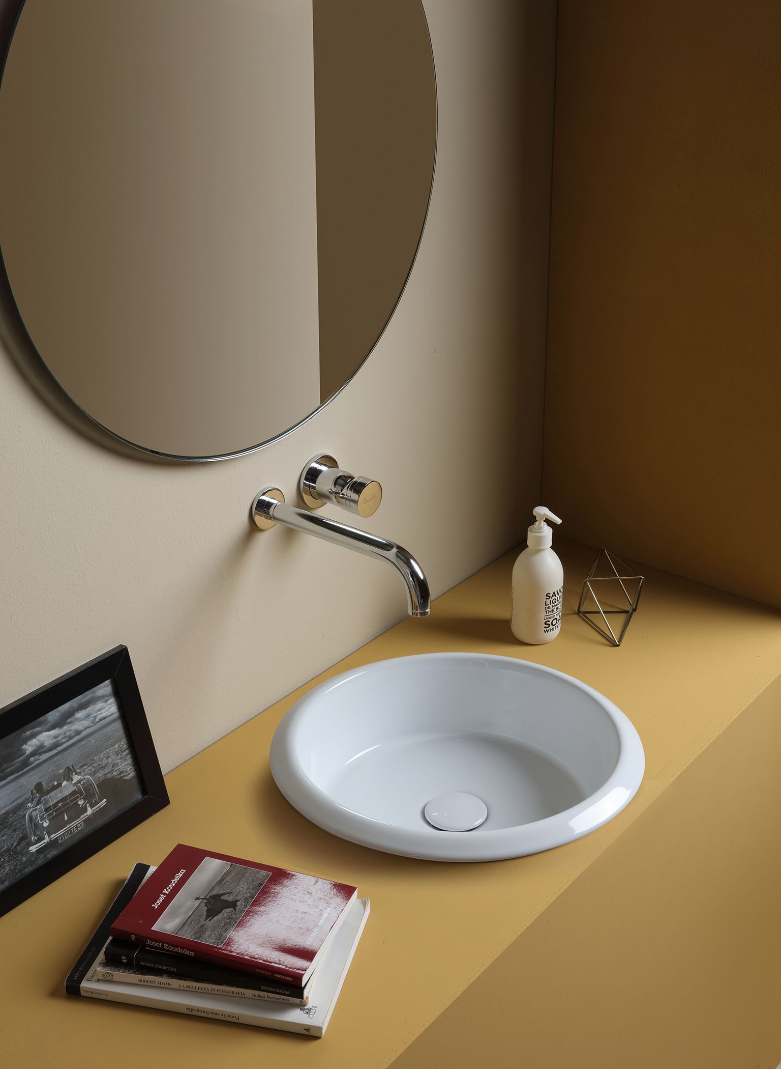 Lavandino, rubinetto e specchio bianchi in un bagno dalle pareti gialle. Libri e sapone sono sul piano di lavoro.