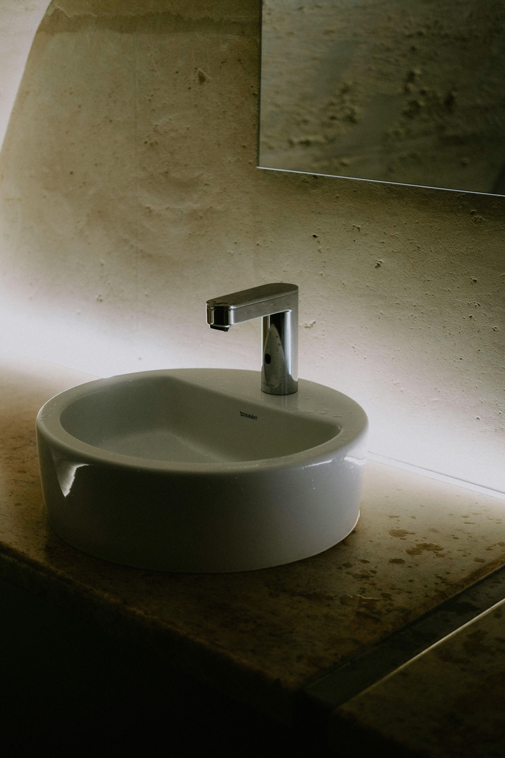 Wall-mounted toilet in a beige-toned bathroom, with a flush panel above. A showerhead is in the background.