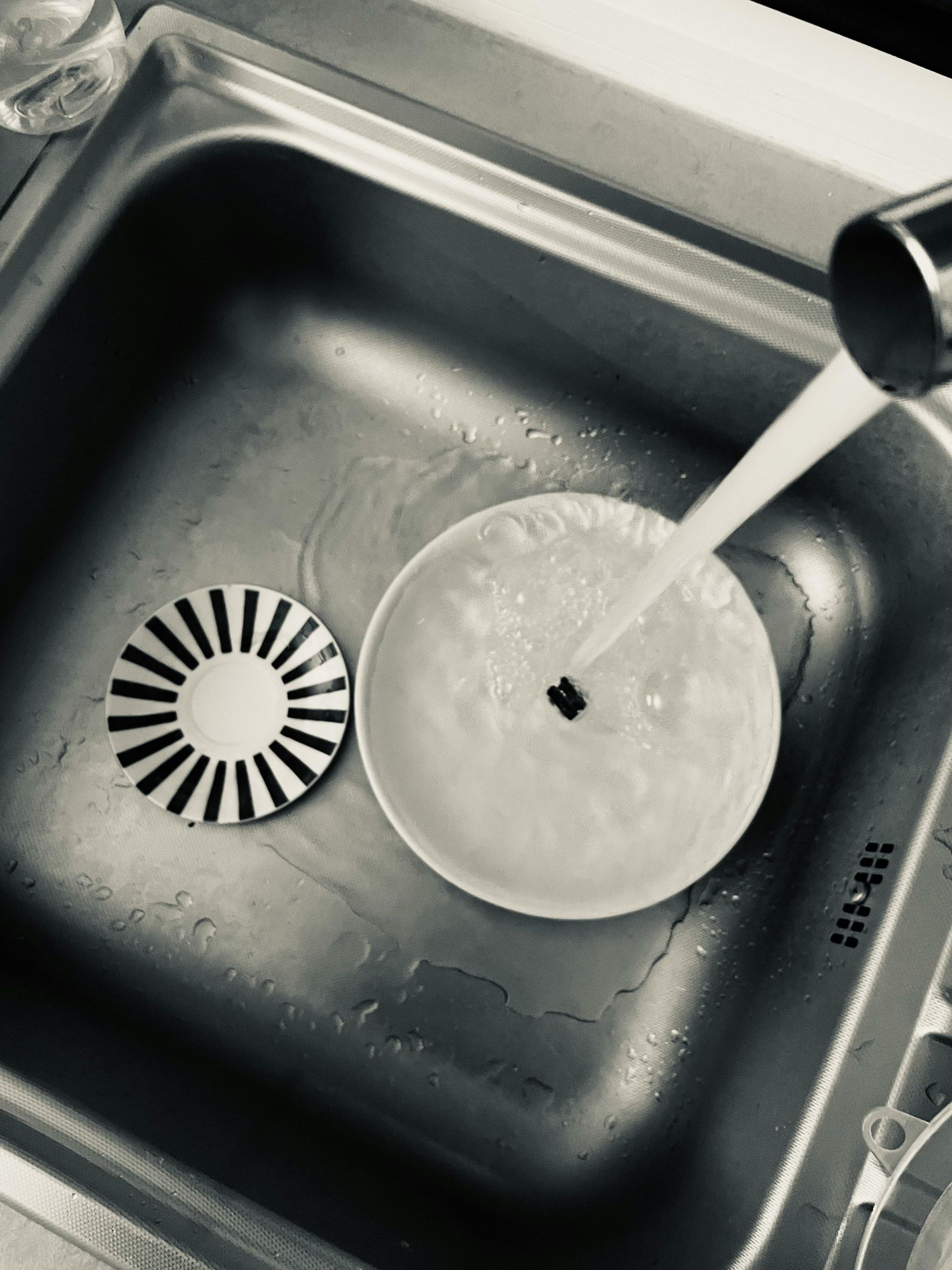 Water flowing onto a white plate in a stainless steel sink, with a striped saucer nearby.