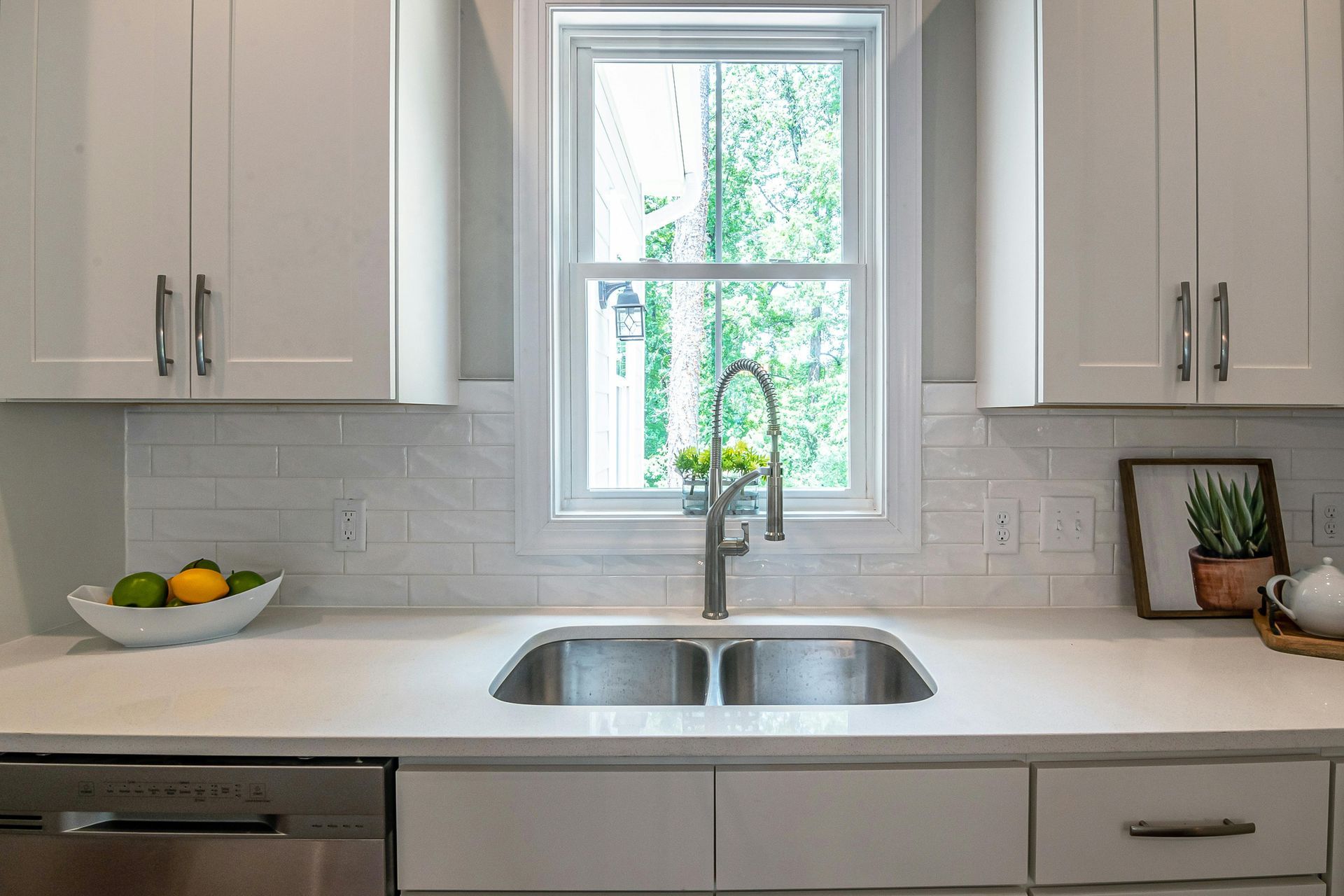 White kitchen with a sink in front of a window, cabinets, and fruit bowl.