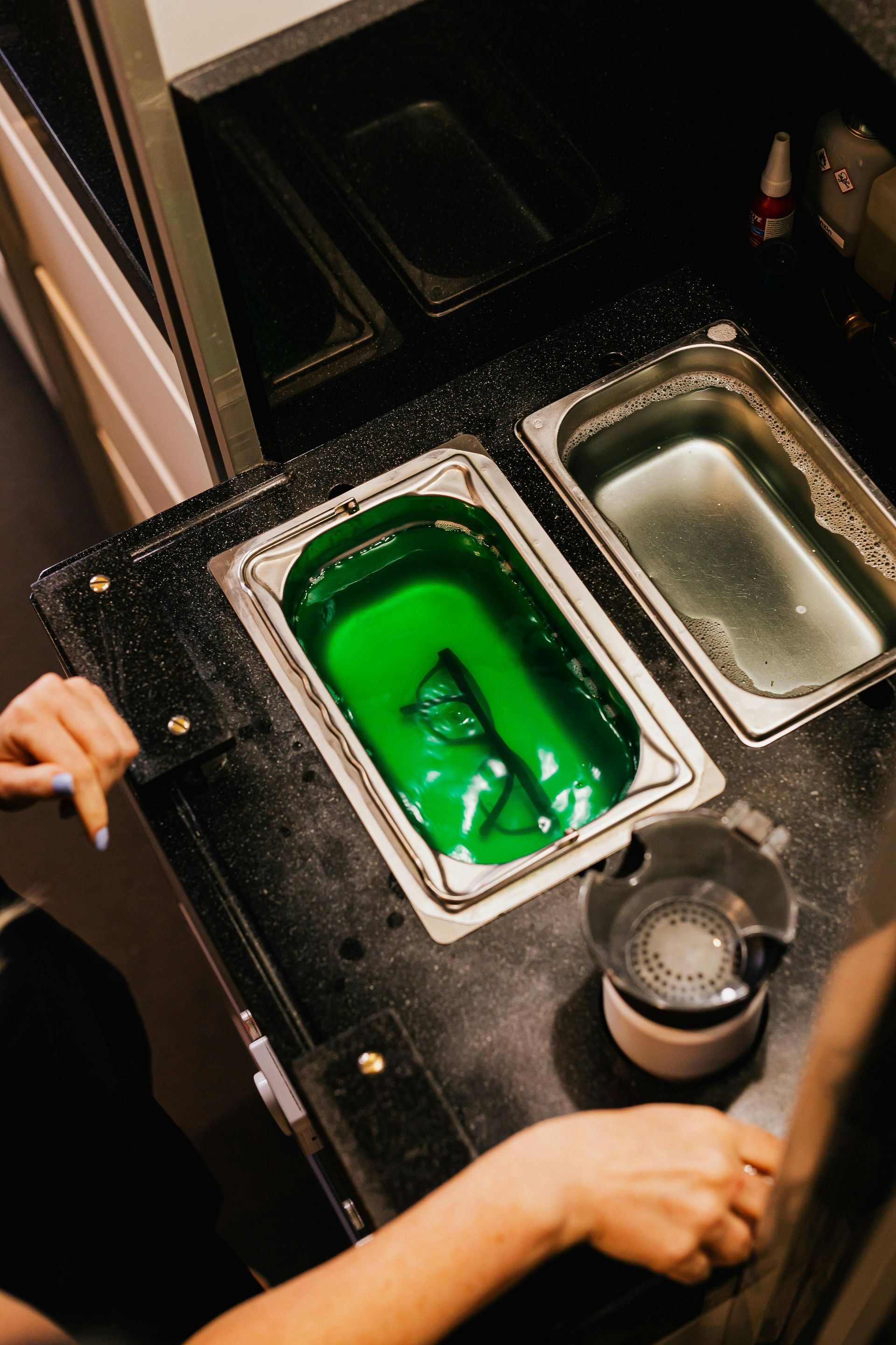 Green dye in metal container on a workstation. Hands reaching towards container and mixing bowl.