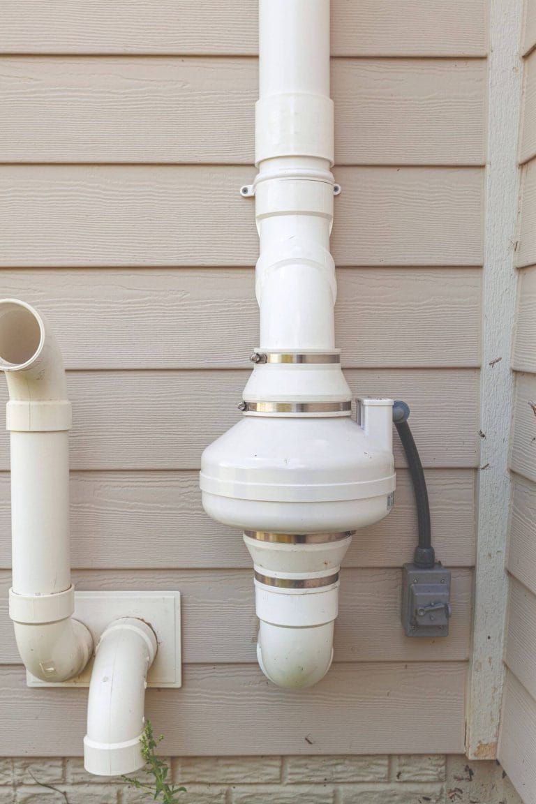 A white radon mitigation system is attached to the side of a house.
