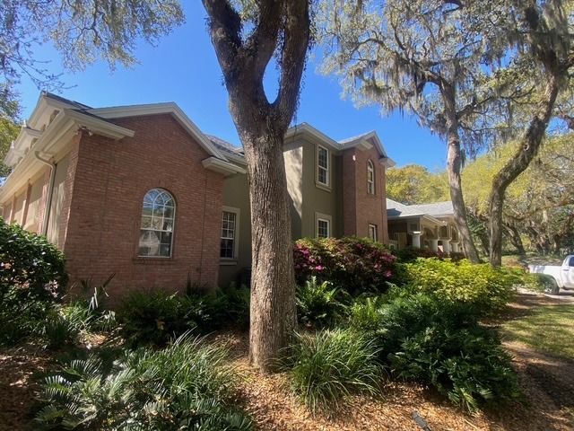 A large brick house is surrounded by trees and bushes on a sunny day.