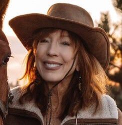 Woman in cowboy hat smiles near a horse, outdoors.