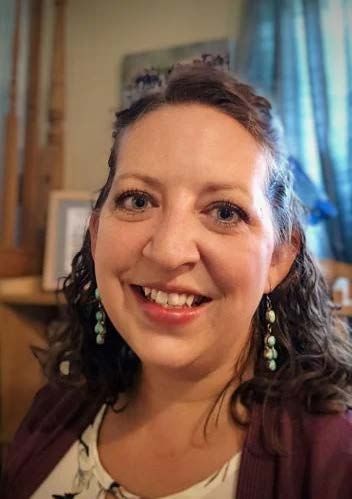Woman with curly brown hair, smiling, wearing teal earrings, indoors.