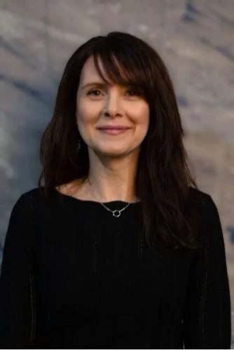 Woman with dark hair and bangs wearing a black top smiles against a blue and gray background.