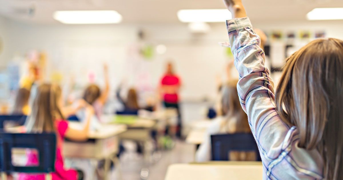 Girl raising her hand in a classroom, other students and teacher blurred in the background.