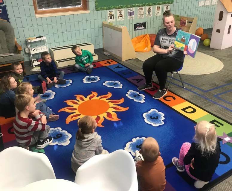 Teacher reading to children seated on a colorful rug. Classroom setting with books and toys.